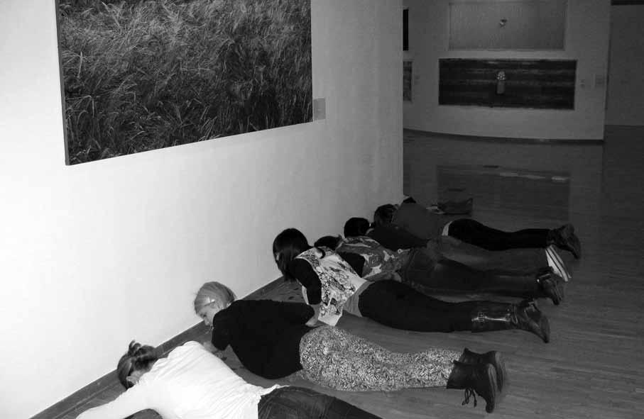 SLIKA 1: Položna fotografija predstavlja prostor v Galeriji Cankarjevega Doma. V diagonali od levega spodnjega do desnega zgornjega dela fotografije je šest oseb, ki ležijo s trupi na parketu.